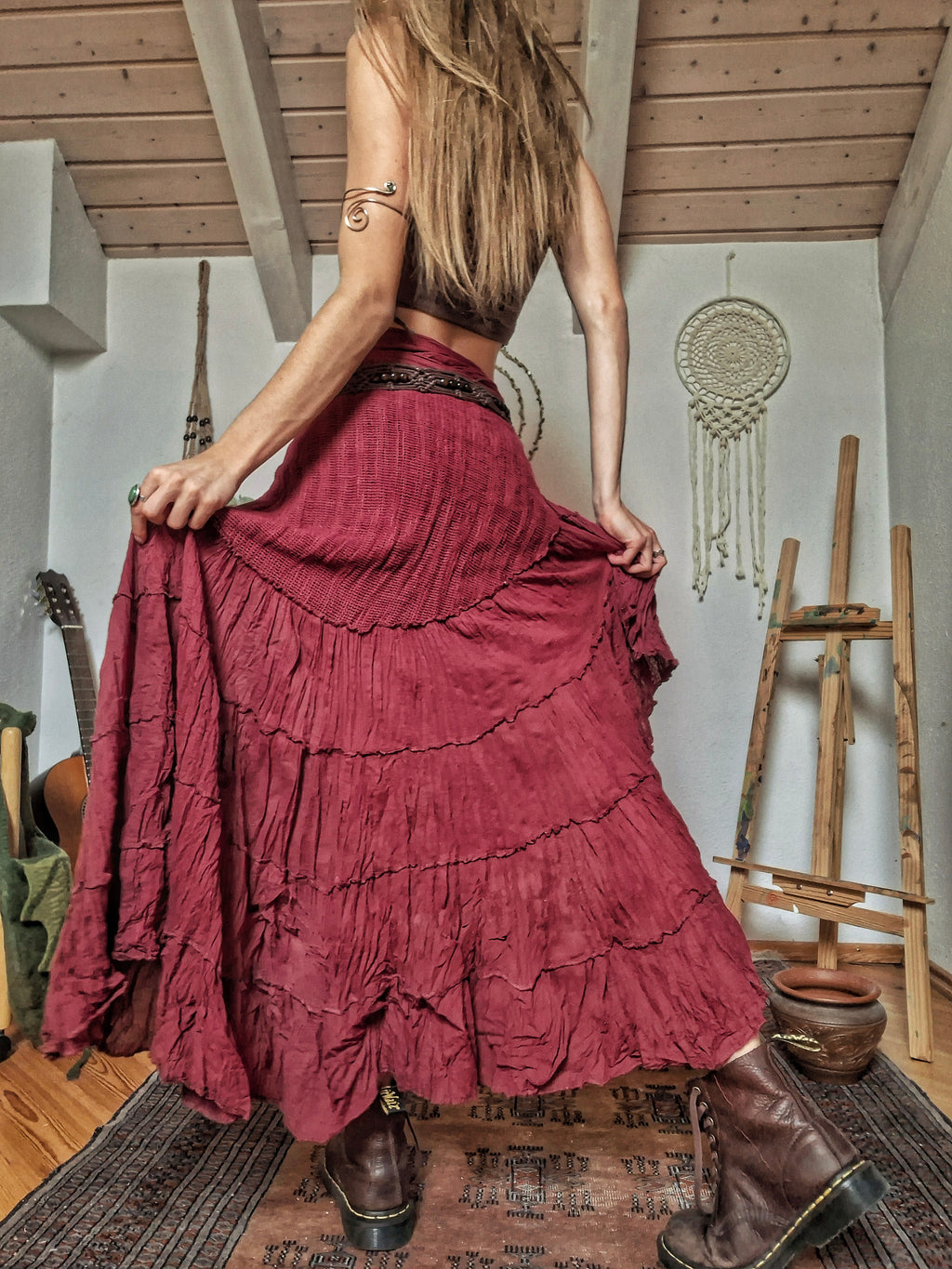 A woman wearing a long, tiered, deep burgundy maxi skirt and brown leather boots, photographed from behind in a rustic room with wooden ceiling beams, a guitar, and a dreamcatcher.