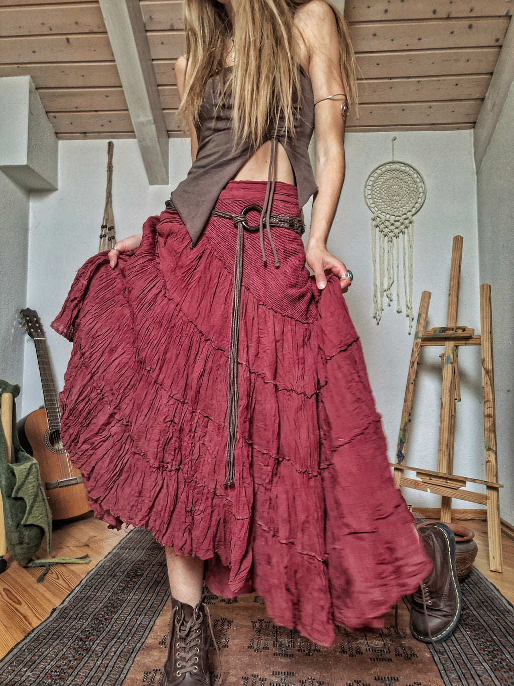 A woman wearing a long, tiered, ruffled maxi skirt in a dusty rose or burgundy red color, paired with a brown cropped top and brown lace-up boots, standing in a rustic room with wooden ceiling beams, a guitar, and an easel.