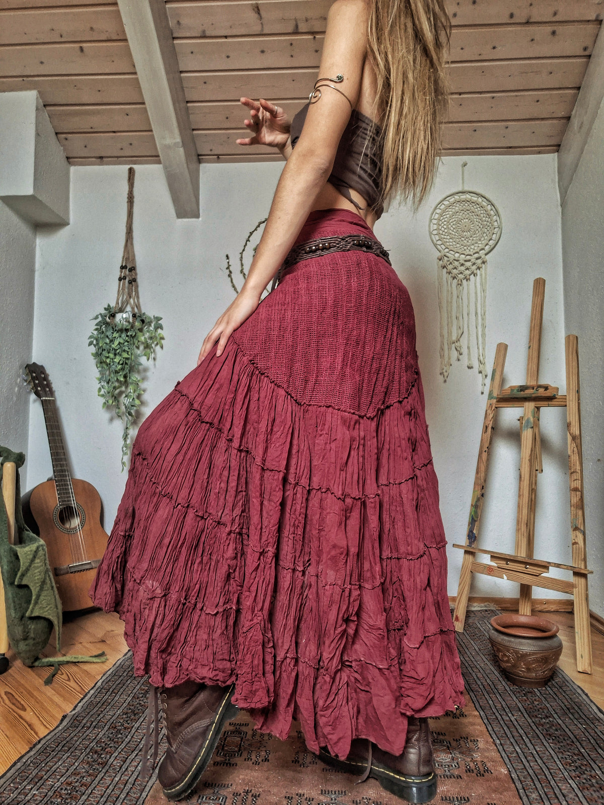 A woman wearing a long, tiered, dusty rose maxi skirt with a dark brown textured top and brown leather boots, standing in a rustic room with a guitar, macrame decor, and wooden ceiling beams.