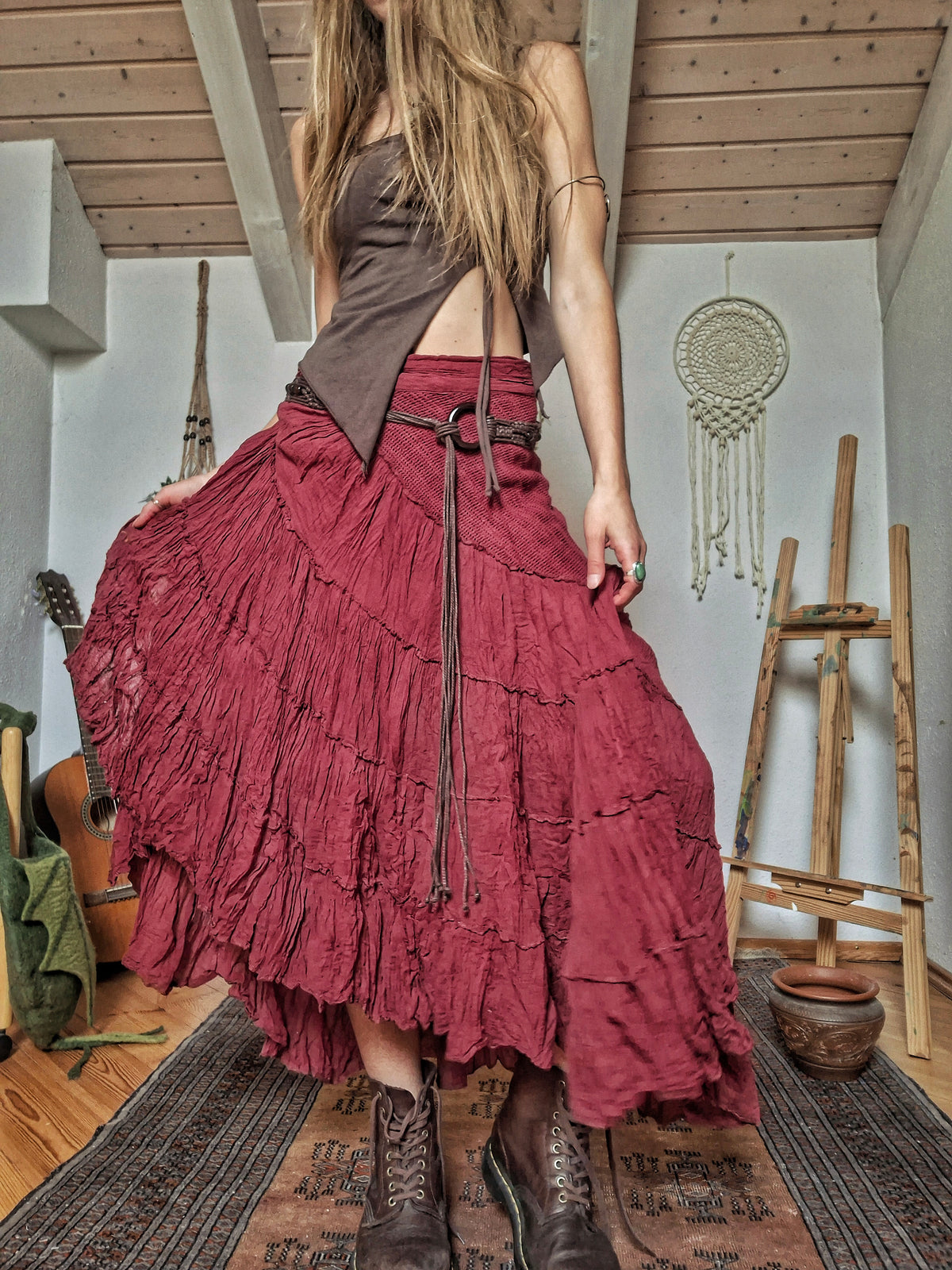 A woman wearing a long, tiered, ruffled maxi skirt in a dusty rose or burgundy shade, styled with a dark brown wrap top and brown lace-up boots, standing in a rustic room with wooden ceiling beams, a guitar, and a dreamcatcher.